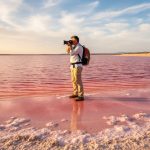 A lone traveler photographing a vivid pink shoreline at Hutt Lagoon, with glowing pink water reflecting golden hour light.