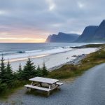 “Dawn coastal view at Gros Morne National Park with ocean waves on a remote white-sand beach and a campground path lined with evergreens toward distant cliffs.”