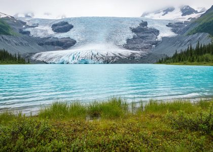 Turquoise glacial lake in Kluane National Park with alpine grass in the foreground and distant mountains and icefield in the background.